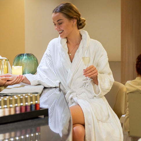 A woman in a white robe sitting in a spa setting, holding a glass of water, with Attendants and other guests in the background.